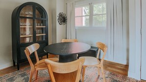 A dining area with a round table, four wooden chairs, a black cabinet with glass doors, and a window with curtains. - Oasis on 5th (Charlottesville)