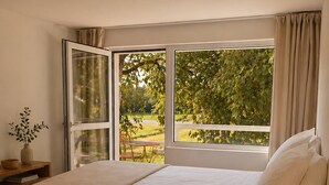 Ein Schlafzimmer mit Bett, Nachttisch mit Lampe, einem Fenster mit Vorhängen und Blick auf Grünfläche draußen.
