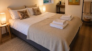 Ein ordentlich bezogenes Bett mit einer beigen Decke und Kissen, ein Nachttisch mit einer Lampe, ein kleines Fenster und ein Bild an der Wand.