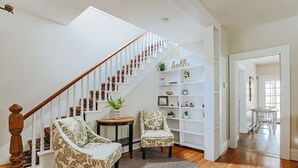 Una escalera con barandilla de madera y paredes blancas que conduce a una habitación con dos sillones estampados, una mesita y un estante blanco con objetos decorativos.