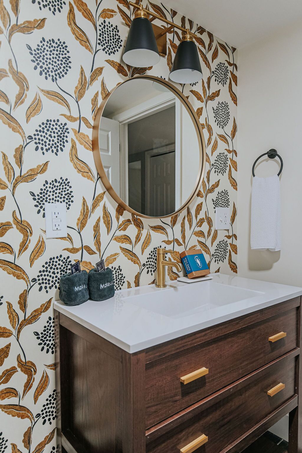 Casa de banho com papel de parede floral, espelho redondo, pia com torneira e porta-toalhas.