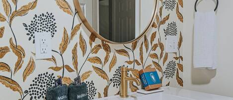 Casa de banho com papel de parede floral, espelho redondo, pia com torneira e porta-toalhas.