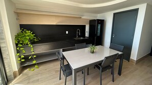 Eine kleine Küche mit schwarzem Arbeitsplatz, integriertem Spülbecken und einem Kühlschrank. Ein Esstisch mit Stühlen und ein Topfpflanze sind ebenfalls vorhanden.