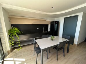 Eine kleine Küche mit schwarzem Arbeitsplatz, integriertem Spülbecken und einem Kühlschrank. Ein Esstisch mit Stühlen und ein Topfpflanze sind ebenfalls vorhanden.