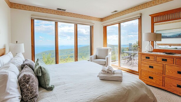 Un dormitorio con una cama grande, un aparador de madera y vistas al paisaje a través de amplios ventanales.