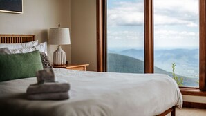Una cama bien hecha con una toalla doblada, una lámpara de noche y vistas a montañas a través de amplios ventanales.