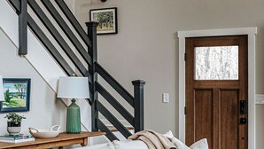 A living room with a white-striped sofa, a wooden coffee table, a green chair, a staircase with black railings, and a wooden door with a window. - Pilotview Hollow (Nellysford)