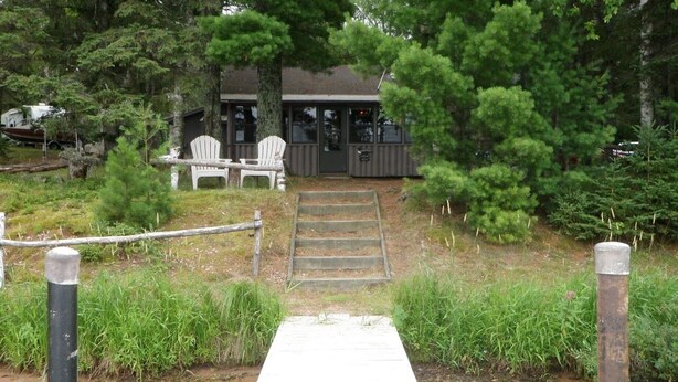 A wooden dock leading to a building with a porch, surrounded by trees and a grassy area. - Charming 3-bedroom lakefront cabin with AC in Hayward Wisconsin! (Hayward)
