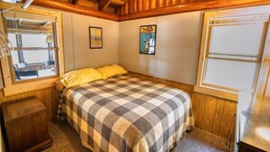 A bedroom with a checkered bedspread, wooden walls, and a window with a view. - Charming 3-bedroom lakefront cabin with AC in Hayward Wisconsin! (Hayward)