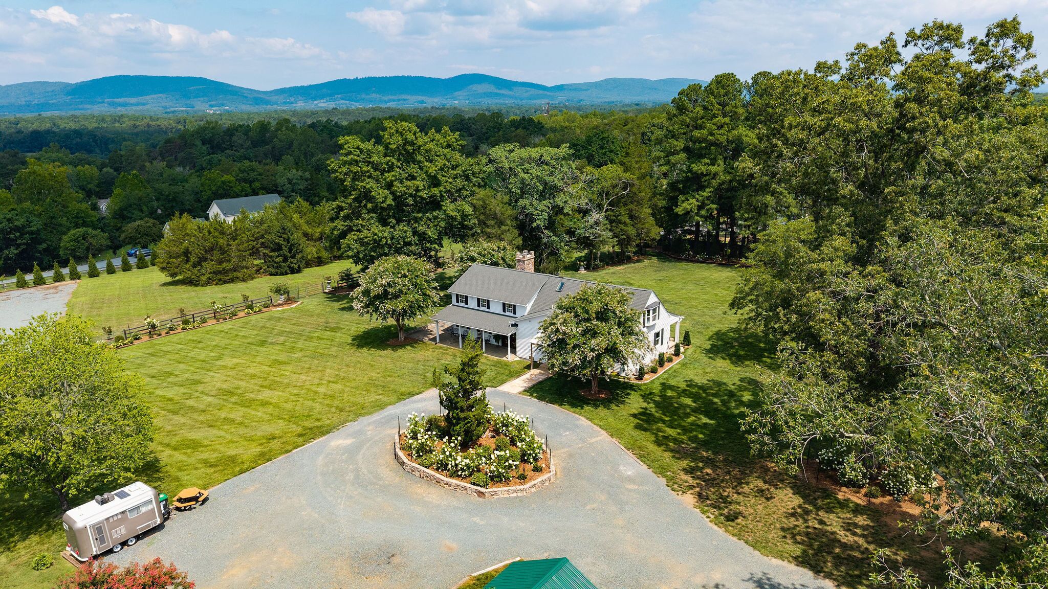 A two-story house with a large lawn, surrounded by trees and hills in the background.