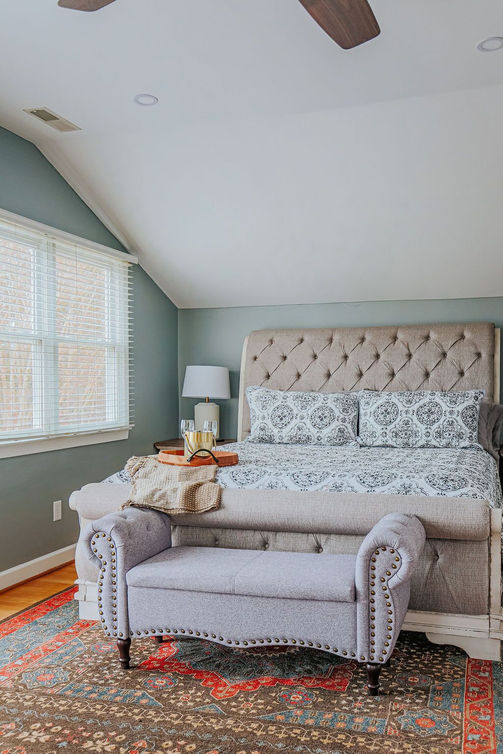 A bedroom with a tufted headboard, a patterned bedspread, a side table with a lamp, a bench, and a patterned rug.