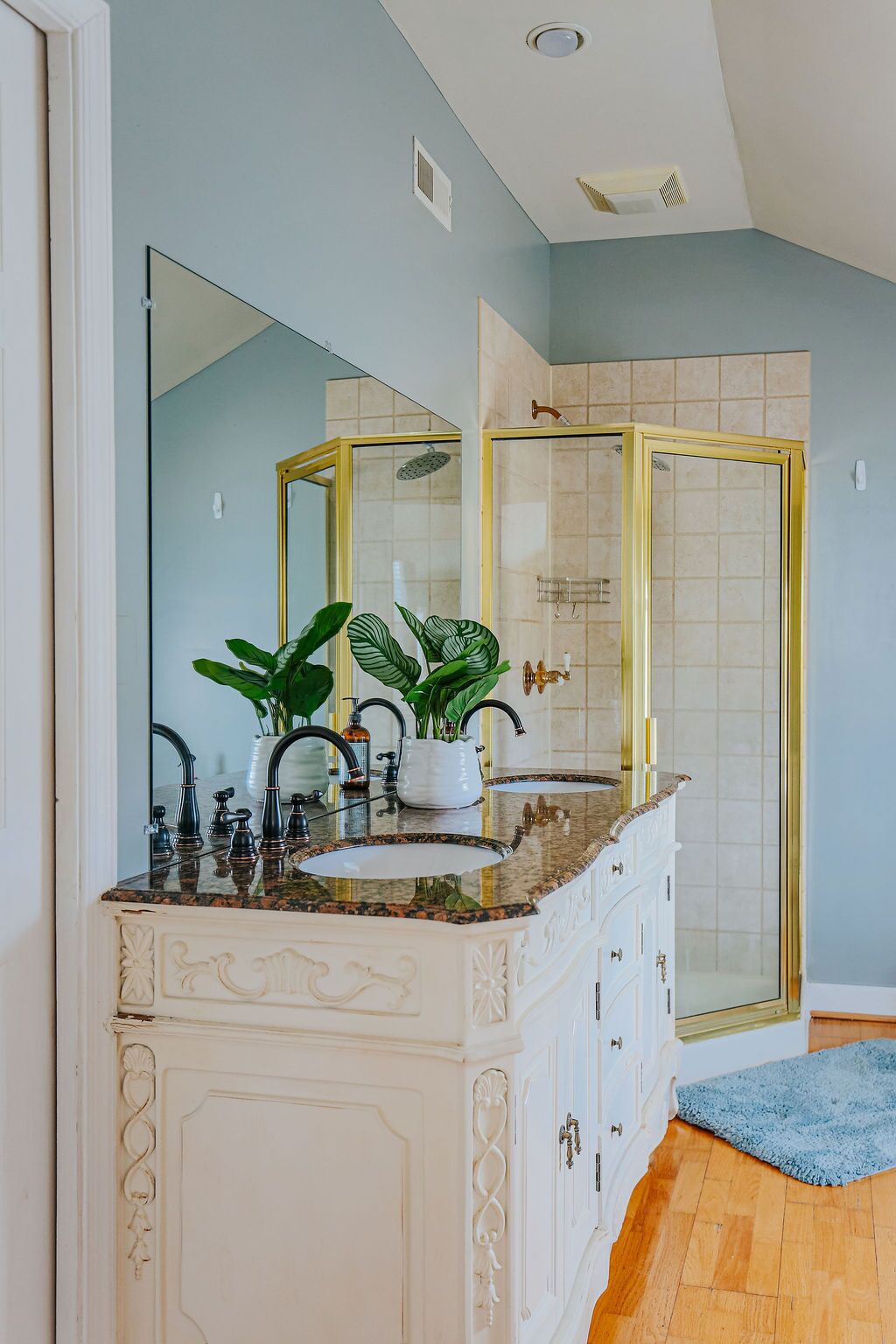 A bathroom with a white vanity, a large mirror, a shower with a gold frame, and a blue rug on the floor.