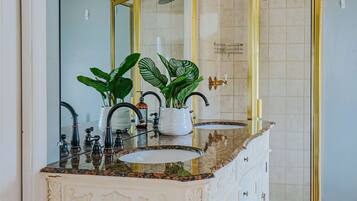 A bathroom with a white vanity, a large mirror, a shower with a gold frame, and a blue rug on the floor.