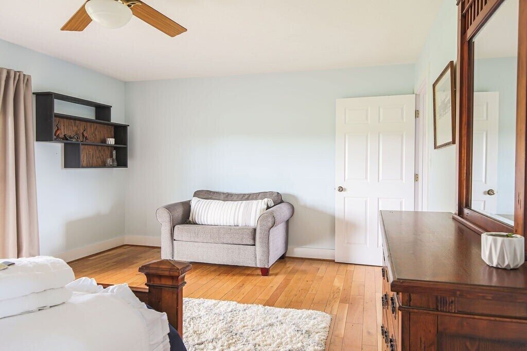 A room with a wooden floor, a grey sofa, a white rug, a wooden dresser, a ceiling fan, and a white door.