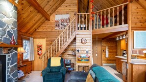 A wooden cabin interior with a fireplace, a staircase, a living room with sofas, and a kitchen area.