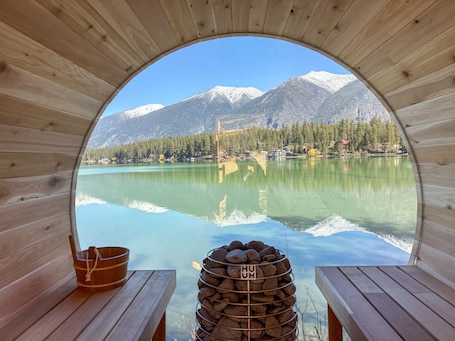 A wooden cabin with a circular window overlooking a calm lake and snow-capped mountains.. Lakefront & Private Dock. A/C. Dog Fr. Rosen Lake