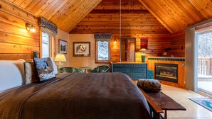 A cozy log cabin room with a bed, a fireplace, a dining table, and a view of the outdoors.