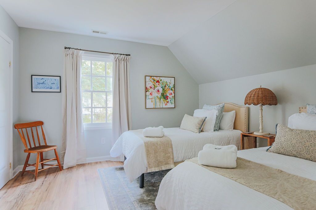 A bedroom with two beds, a wooden chair, a window with curtains, a lamp, and a framed picture on the wall.