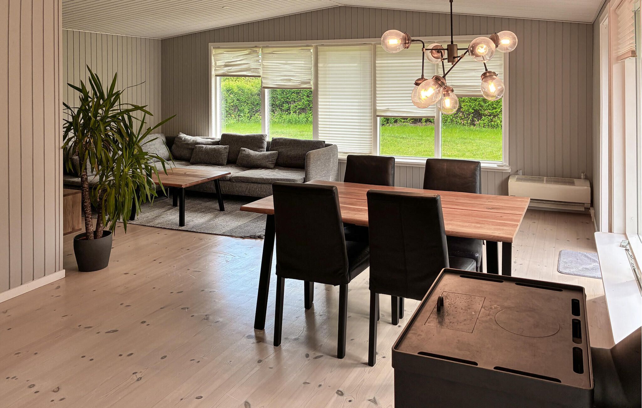 A dining area with a wooden table, black chairs, a potted plant, and a large window with blinds.