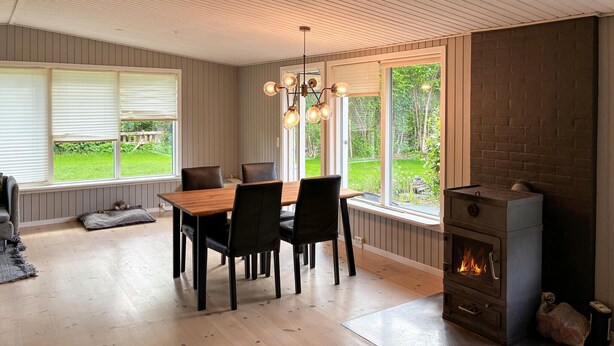 Un espace repas comprenant une table et des chaises en bois, une cheminée et de grandes fenêtres donnant sur un paysage verdoyant.