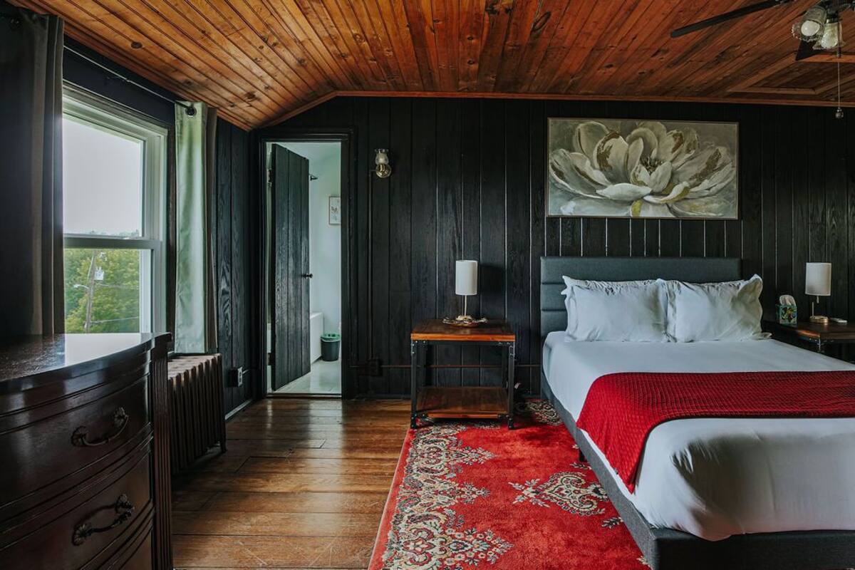 A bedroom with a wooden ceiling, a large bed with white bedding, a red rug, and a wall-mounted flower painting.