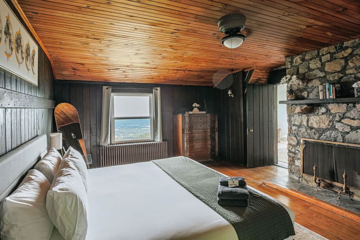 A bedroom with a wooden ceiling, a stone fireplace, a bed with white linens, and a view through a window.
