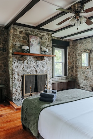 Un dormitorio con pared de piedra, una chimenea, una cama con manta verde y un ventilador de techo.