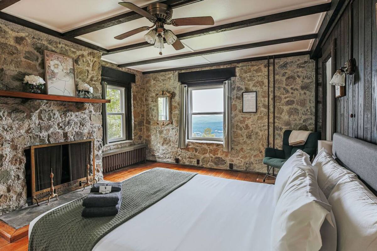 A bedroom with a stone wall, a bed, a fireplace, and a view of the sea.