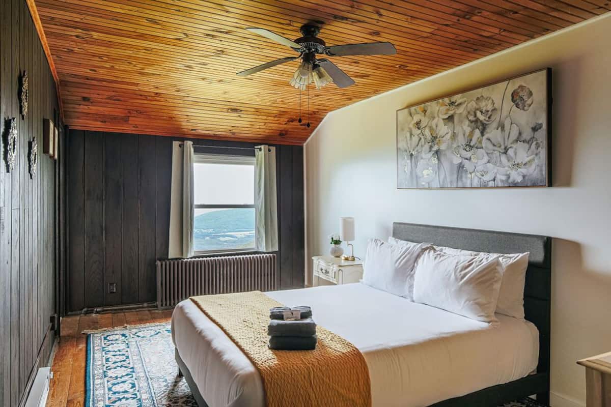 A bedroom with a wooden ceiling, a bed with white bedding, a blue patterned rug, a ceiling fan, a window with a view, and a painting on the wall.