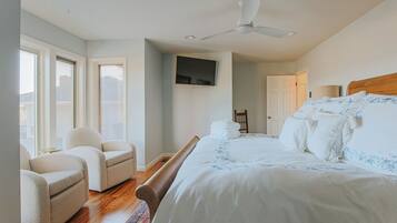 Un dormitorio con cama, dos sillones, ventilador de techo, televisor montado en la pared y una puerta que da a otra habitación.