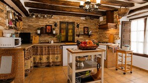 A rustic kitchen with stone walls, wooden beams, a tiled floor, a central island with a fruit bowl, and a hanging light fixture. - Casa rural de gran capacidad (Alcala la Real)