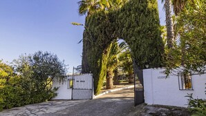 Una entrada con portón y un camino que conduce a un edificio, rodeado de árboles y arbustos.