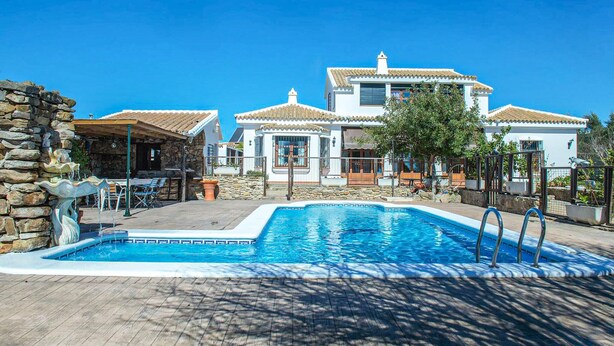 A pool area with a stone wall, a wooden deck, and a large house with a tiled roof.