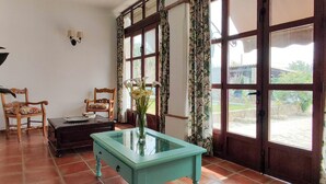 A room with a glass-top table, two chairs, a small table with a vase of flowers, and large windows with curtains.