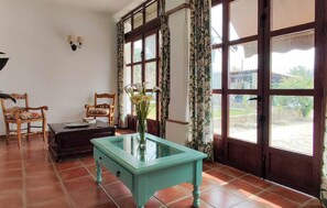 Ein Zimmer mit Glastisch, zwei Stühlen, einem kleinen Tisch mit Blumenvase und großen, mit Vorhängen versehenen Fenstern.