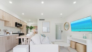 A modern kitchen with white cabinets, a dining area with a white table and bench, a flat-screen TV mounted on the wall, and a view of the sea through the window.