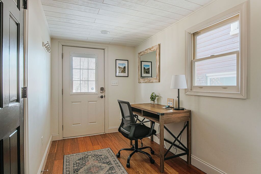 A room with a wooden desk, black office chair, and a small table with a lamp. There is a door with a window and a mirror on the wall.