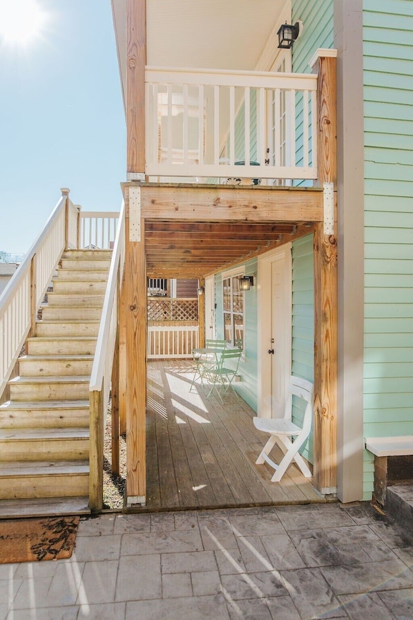 A wooden deck with a staircase leading to a covered area, a small outdoor seating area with chairs, and a view of a house with light green siding. - The Hunker Down (Staunton)