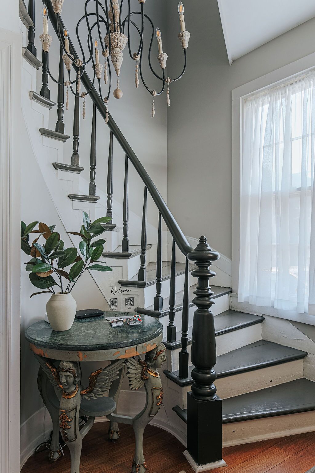 A staircase with black balusters and a chandelier, a round table with a vase and a QR code, and a window with sheer curtains.