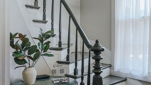 Uma escadaria com balaústres pretos e um candelabro, uma mesa redonda com um vaso e um código QR, e uma janela com cortinas leves.