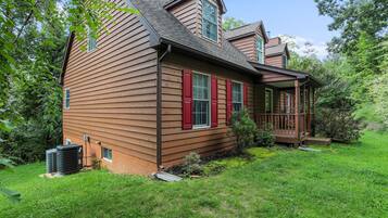A two-story wooden house with a covered porch, a small garden, and an air conditioning unit.