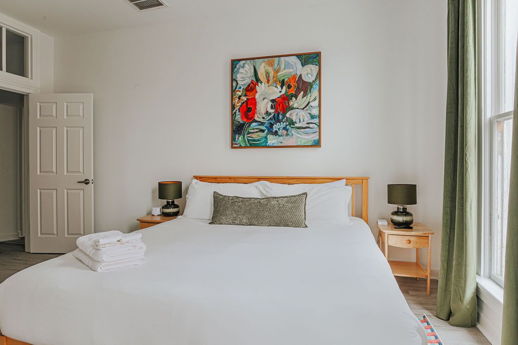 Une chambre d’hôtel avec un grand lit, deux tables de chevet avec des lampes, un tableau au mur et une porte sur le côté gauche.