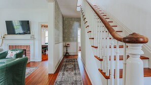 Eine Treppe mit weißen Balustern und einem hölzernen Geländer führt zu einer oberen Etage. Im Vordergrund sind ein grüner Sessel und ein Kamin zu sehen.