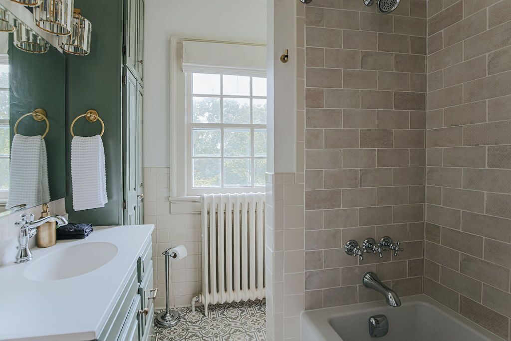Un baño con un lavamanos blanco, un espejo, un perchero con toallas blancas, una ventana, un radiador y una zona de ducha con azulejos color beige.