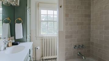 Un baño con un lavamanos blanco, un espejo, un perchero con toallas blancas, una ventana, un radiador y una zona de ducha con azulejos color beige.