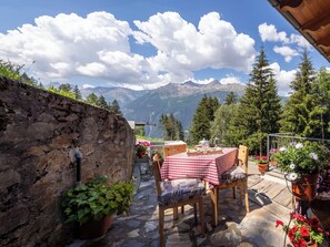 A stone wall, a table with a checkered tablecloth, chairs, potted plants, and a mountainous landscape in the background. - Chalet Ralon by Interhome (Bormio)