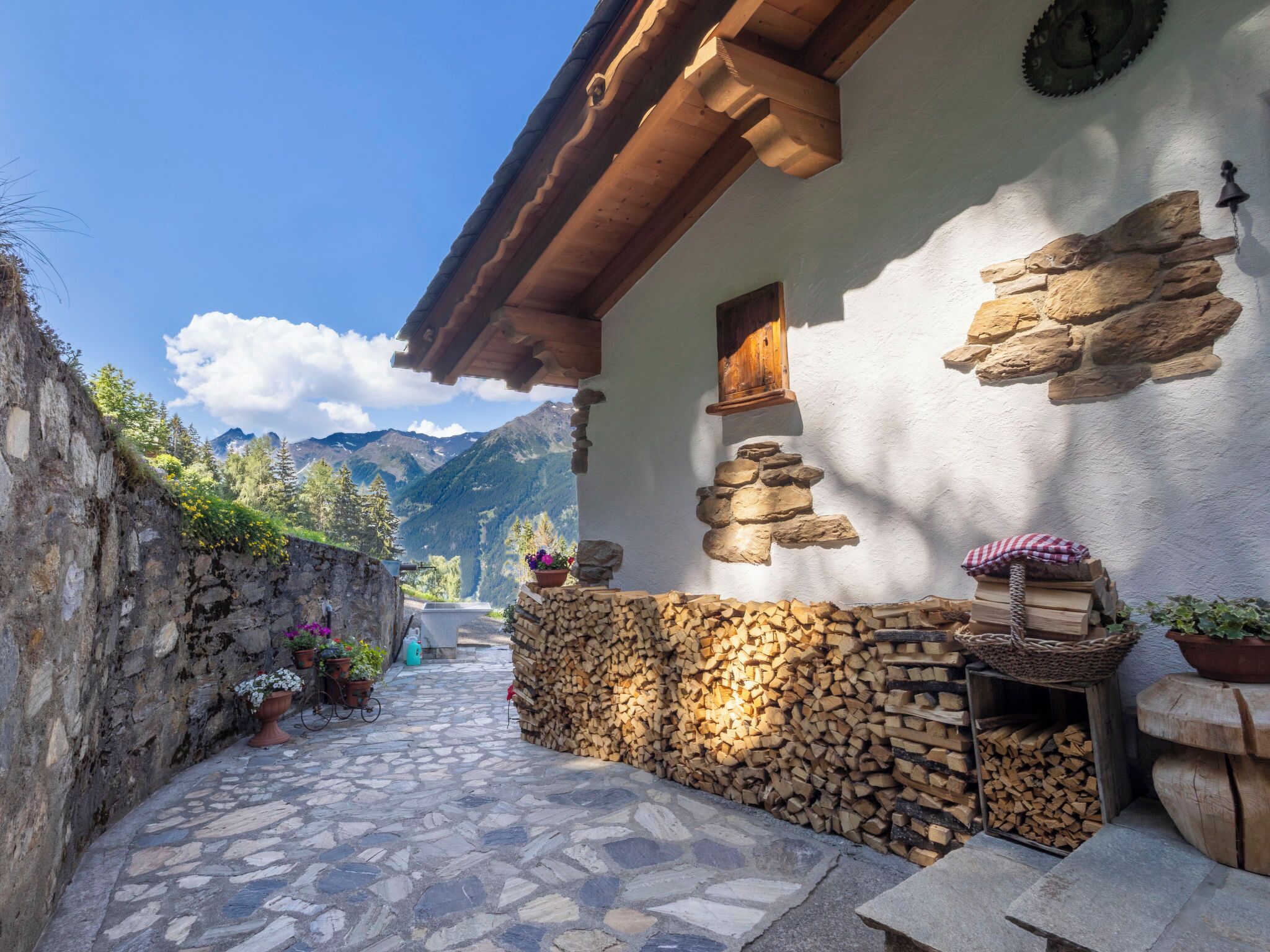 Une cour dallée de pierres, avec une structure en poutres de bois, un mur en pierre et une vue sur les montagnes.