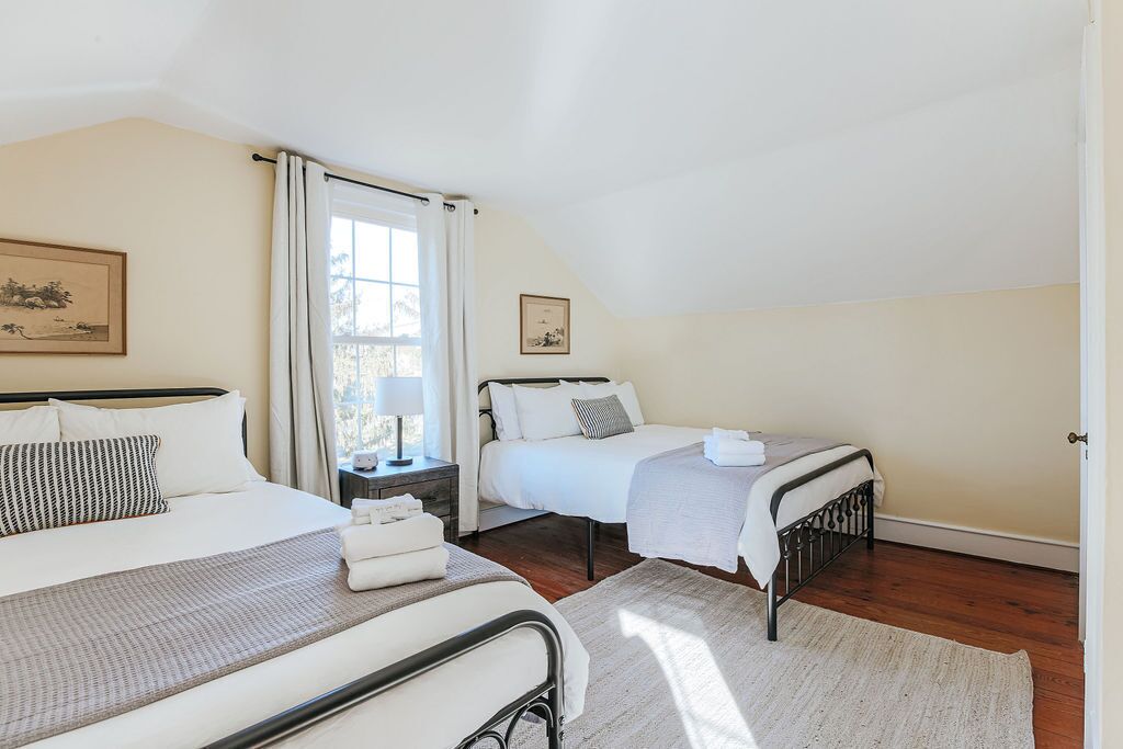 Une chambre avec deux lits, une table de chevet, une fenêtre avec des rideaux et un tableau encadré au mur.