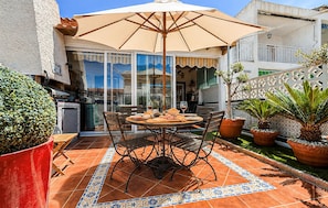 Eine Terrasse mit Tisch und Stühlen, einem Sonnenschirm, Blumentöpfen und Blick auf die Straße.