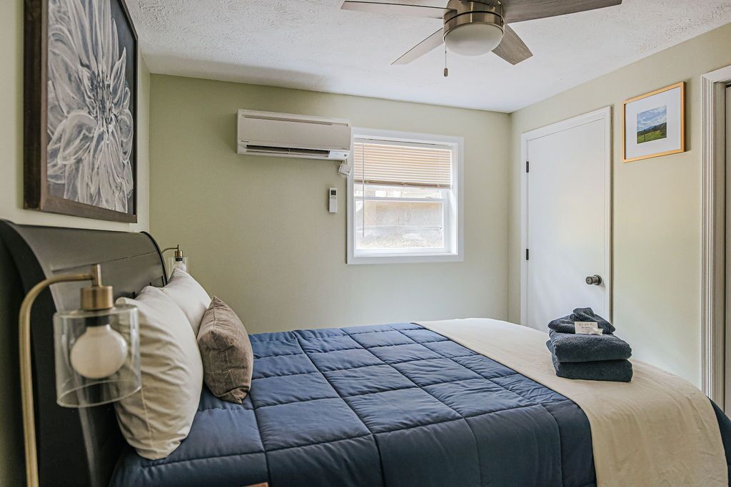 Une chambre à coucher comprenant un lit, un ventilateur de plafond, un climatiseur, une fenêtre avec des stores et une porte.